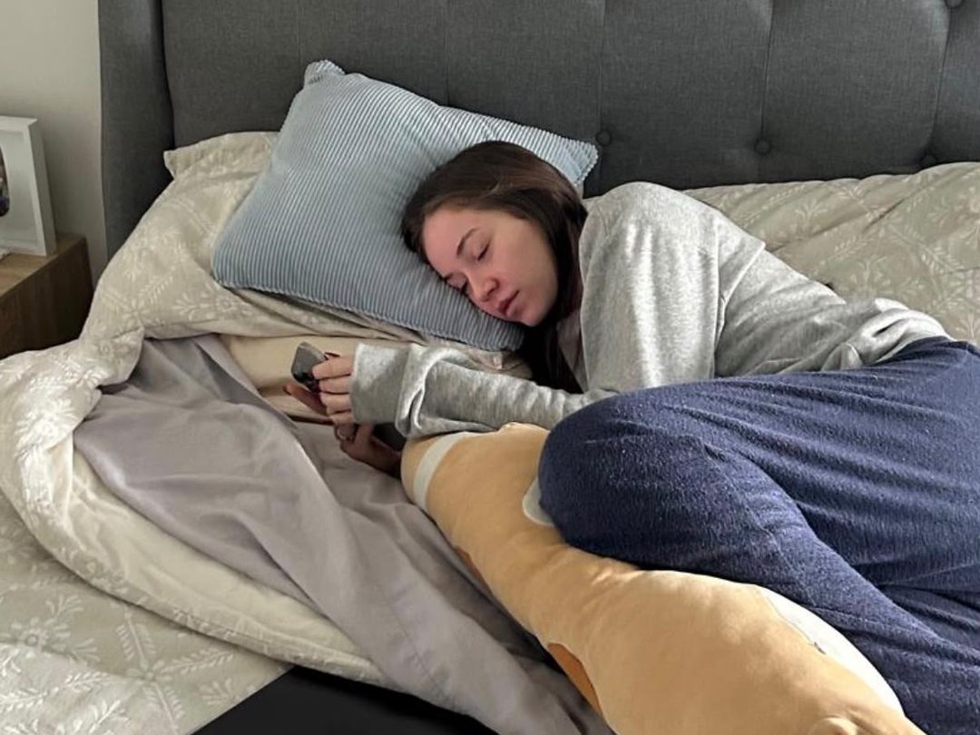 Vanessa asleep on top of a made bed in the daytime, her phone still in her hand.